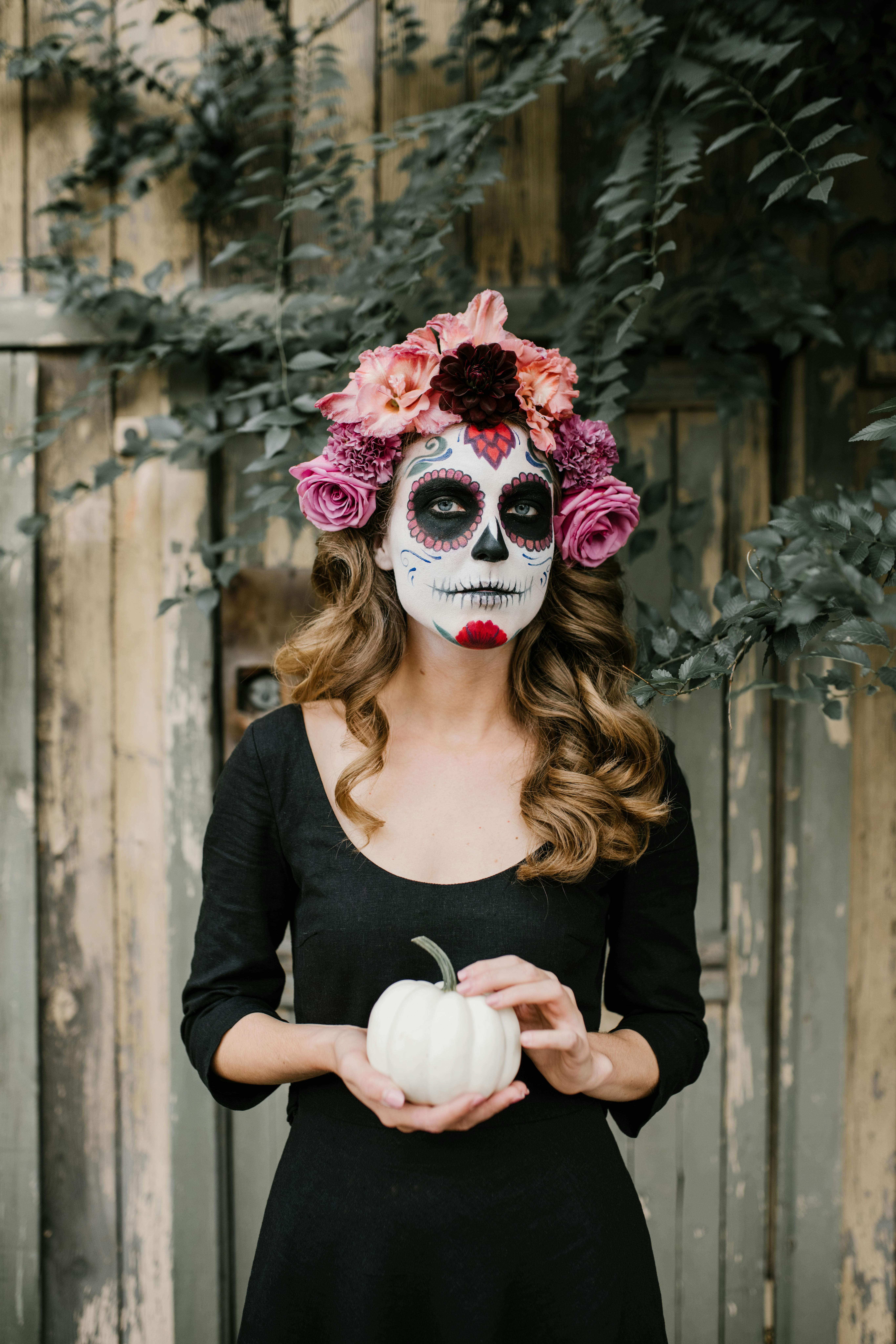 Woman with Day of the Dead makeup holding a white pumpkin and wearing a flower crown.