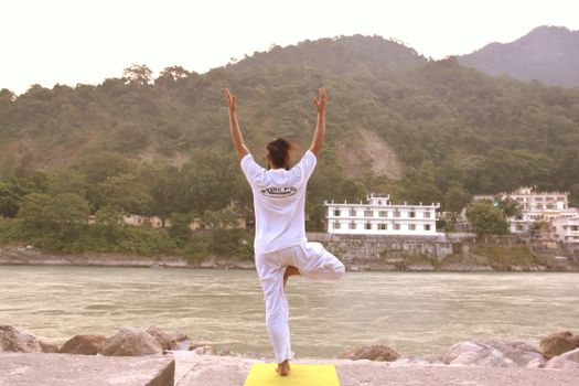 Back view of a man in a yoga pose by the Ganges River, Rishikesh, India, amidst serene nature.