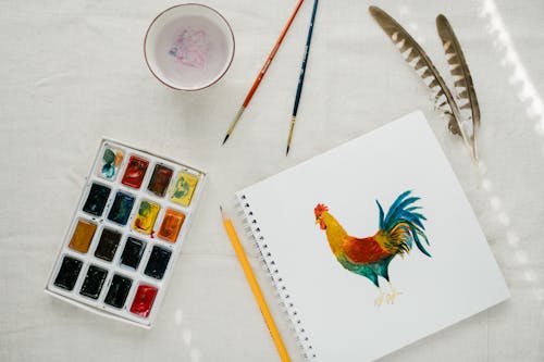 From above of creative composition of sketchbook with painted cock set of watercolors paintbrushes and