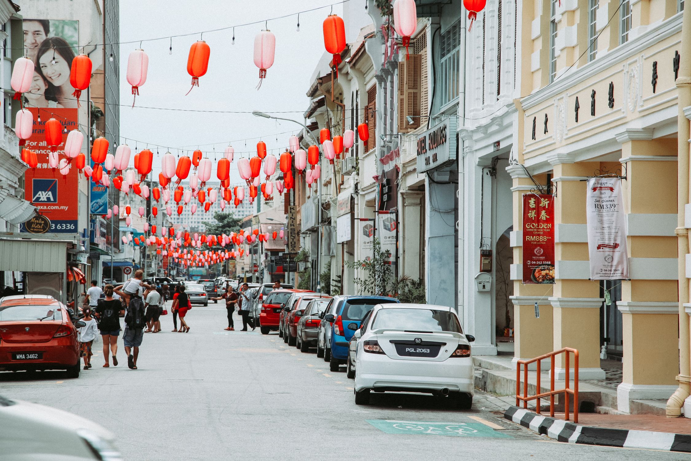 Crowd of people strolling on asphalt road with parked cars on street with residential buildings and hanging Chinese lanterns in city
