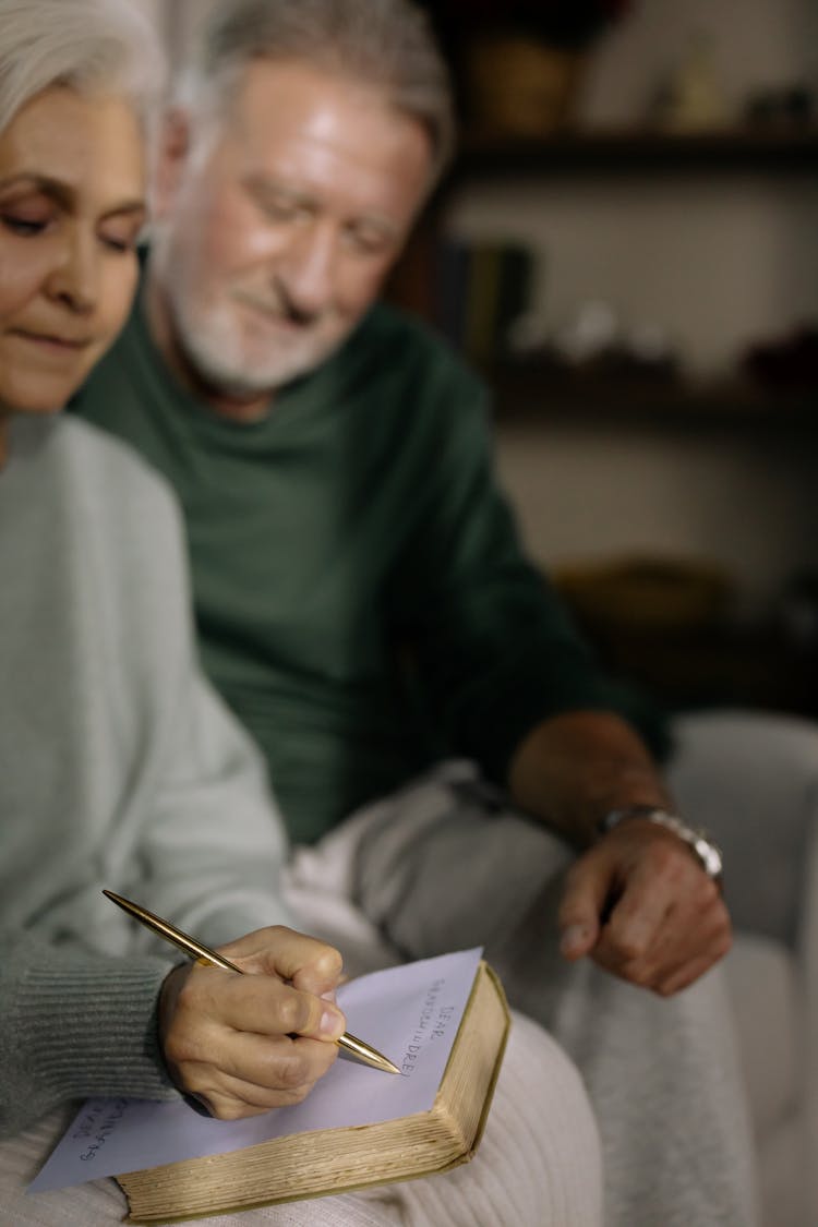 Elderly Woman Sitting Beside Her Husband Writing On A Paper 