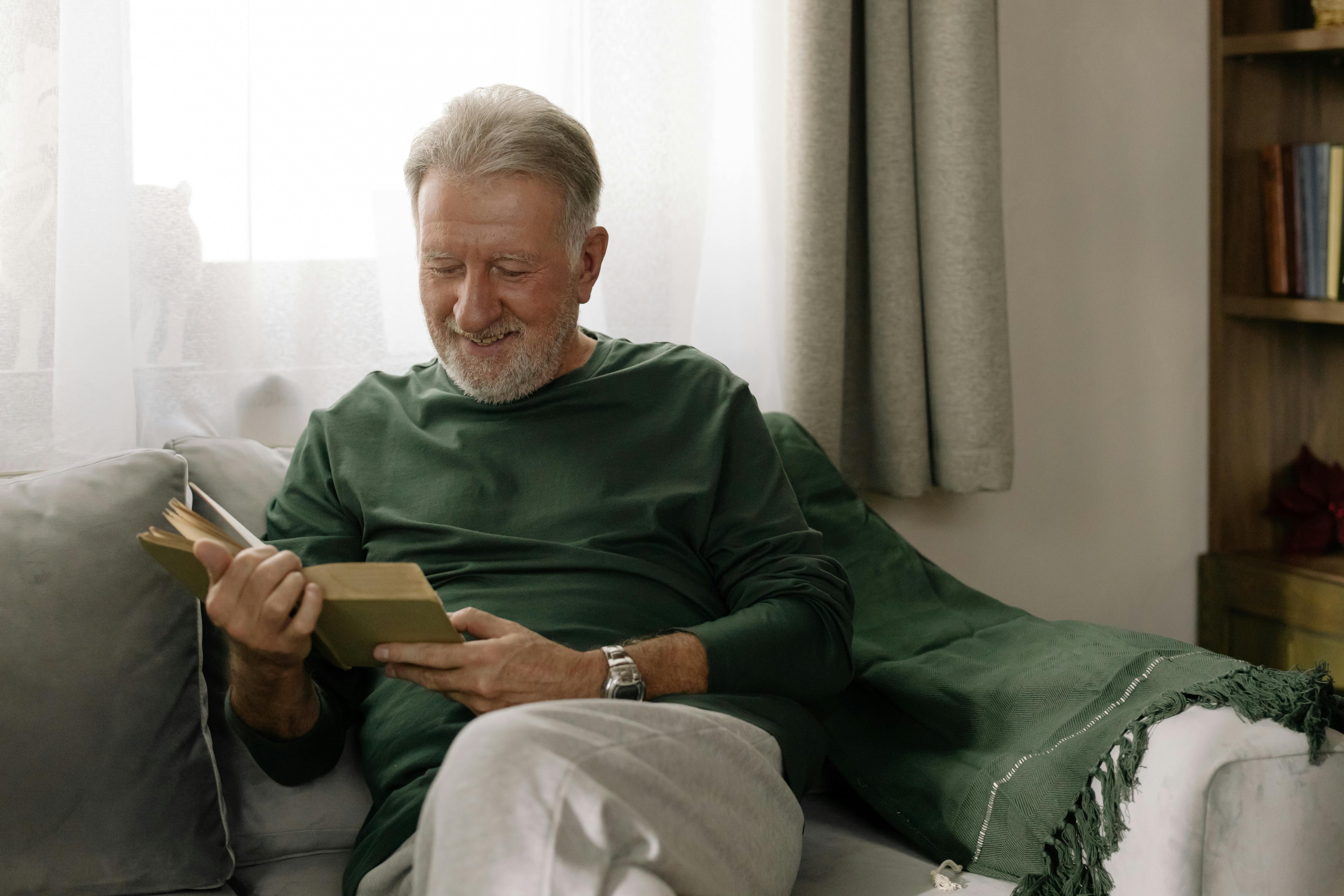 Elderly man with a beard reading a book on a sofa, expressing joy and relaxation in a cozy living room setting.