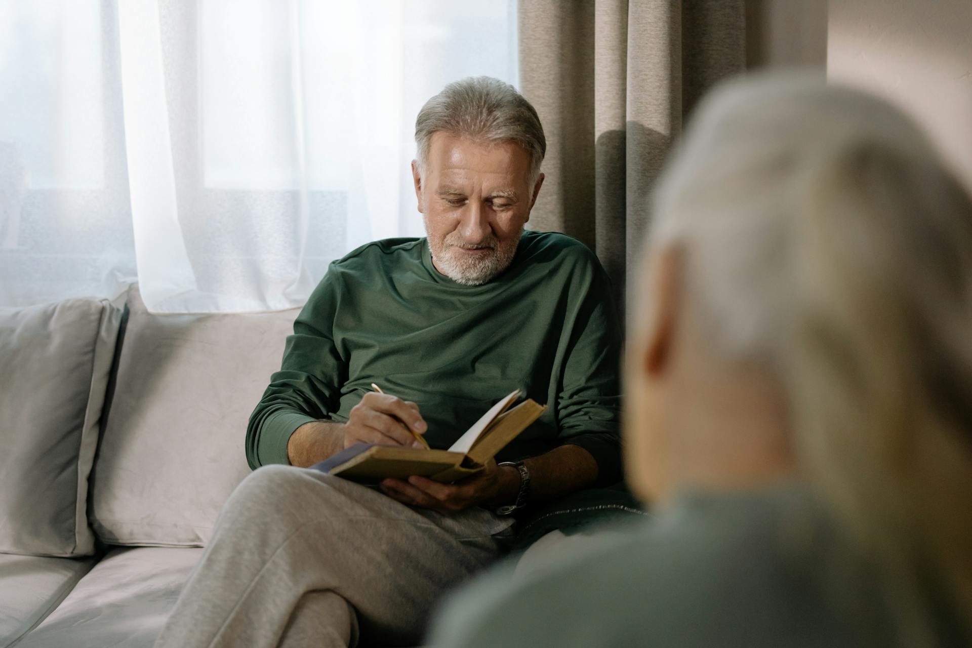 Senior man in a green sweater reading a book on a sofa, enjoying a peaceful indoor setting.