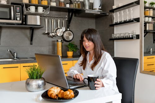 Woman in White Long Sleeve Shirt Sitting at the Table Using a Laptop 