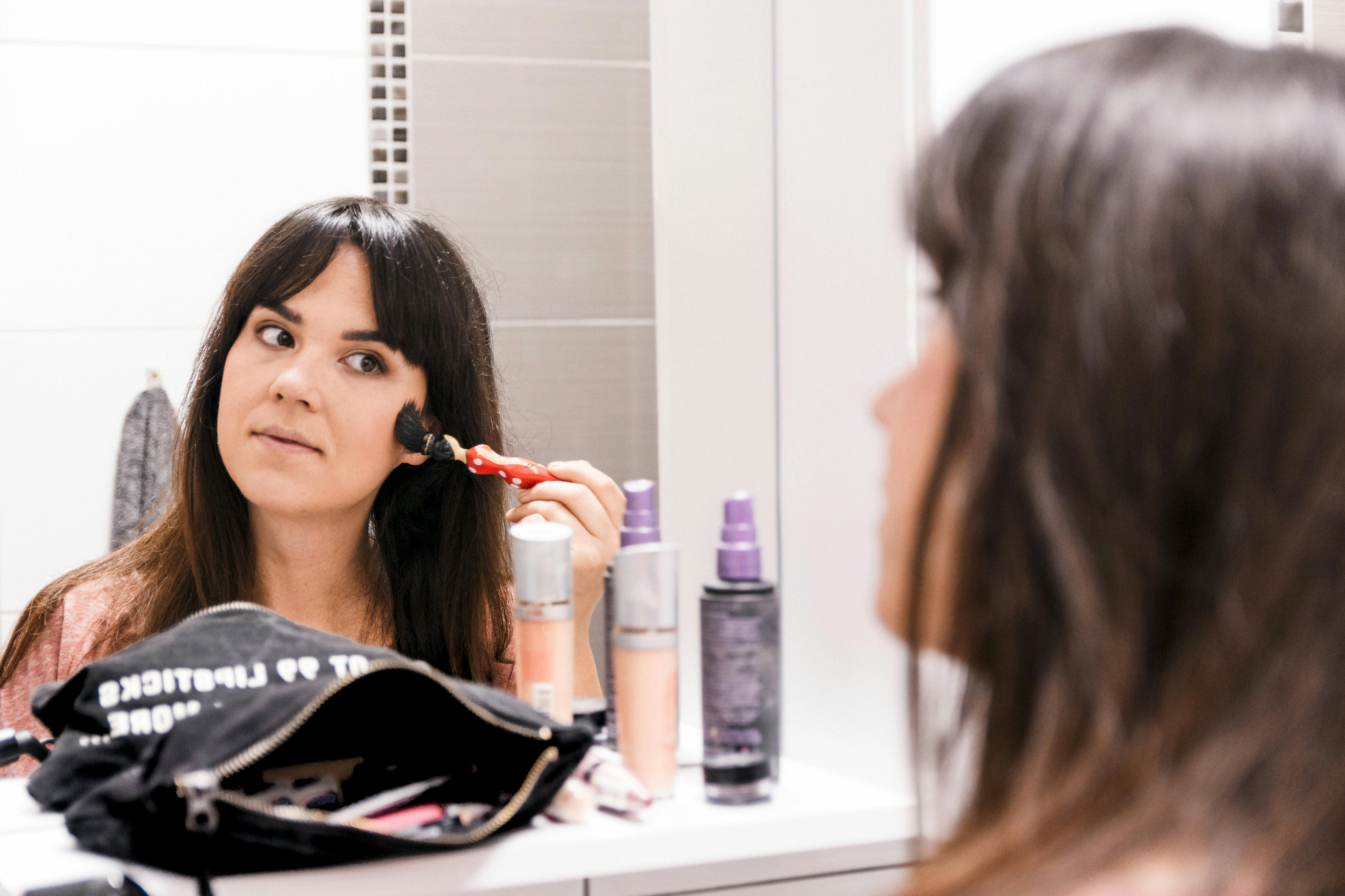 A woman applying makeup using a brush in front of a bathroom mirror, surrounded by cosmetics.