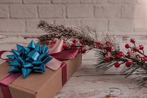 High angle of carton gift box tied with ribbon bow placed on wooden table near Christmas tree branch with snowflakes and red berries