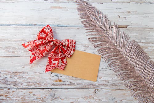 Top view of red and white bow with Christmas pattern and small kraft card placed on wooden table with decorative glitter feather