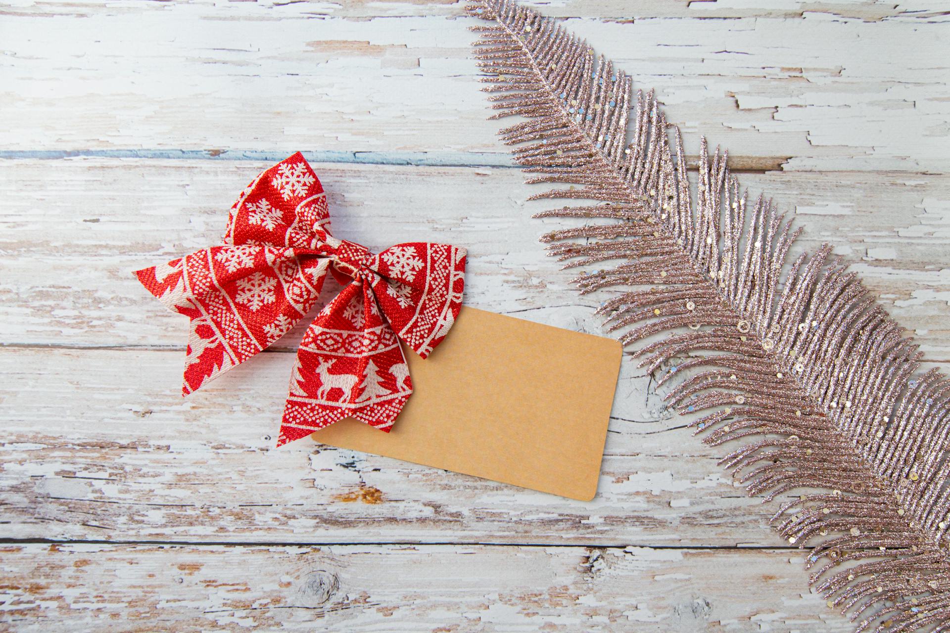 Top view of red and white bow with Christmas pattern and small kraft card placed on wooden table with decorative glitter feather