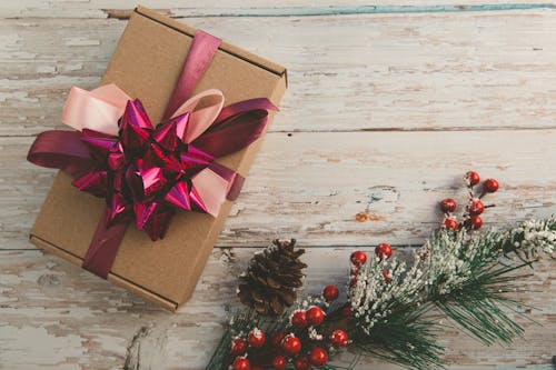 From above elegant gift box with ribbon bows placed on rough wooden table near branch of green Christmas tree with red berries and cone