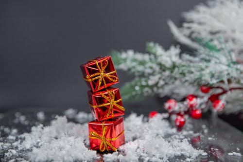Small gift boxes wrapped in shiny red paper and tied with golden bows stacked on table covered with snow near Christmas tree against gray wall