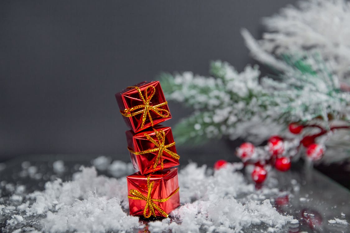 Free Small gift boxes wrapped in shiny red paper and tied with golden bows stacked on table covered with snow near Christmas tree against gray wall Stock Photo
