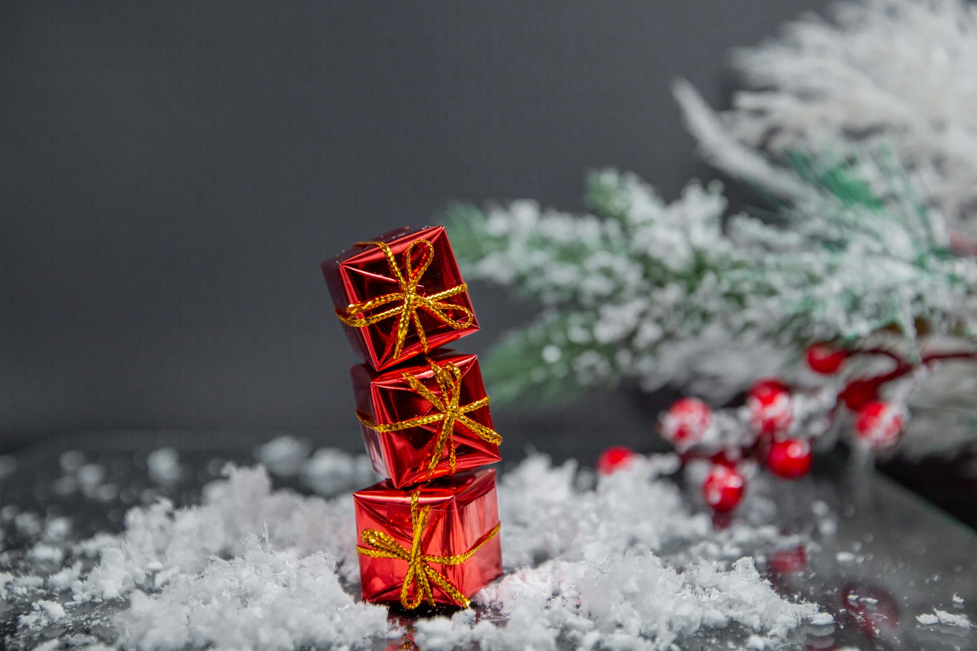 Small gift boxes wrapped in shiny red paper and tied with golden bows stacked on table covered with snow near Christmas tree against gray wall