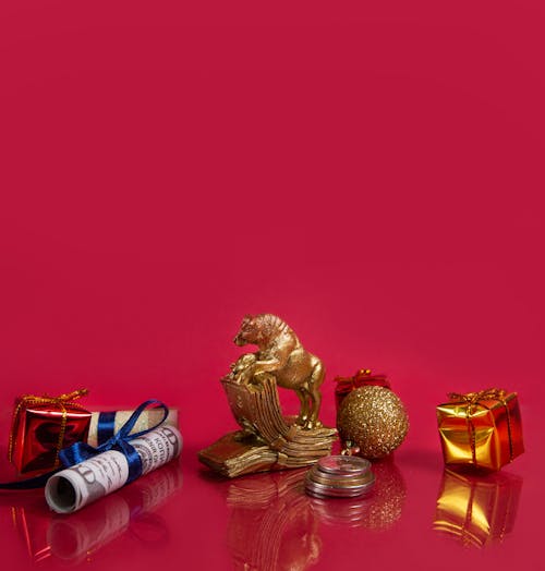 Symbolic golden bull figurine with wrapped New Year gifts and bauble placed on shiny table with coins stack and rolled dollar banknotes against red background