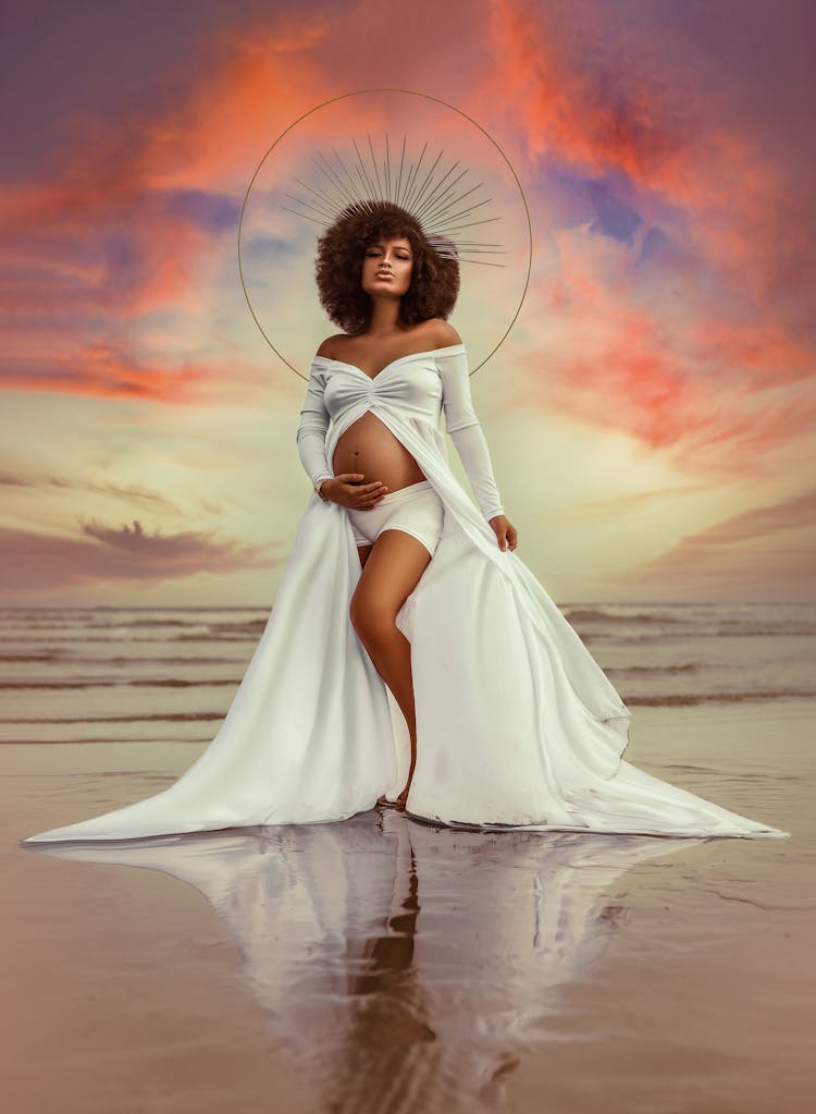 Black Pregnant Woman With Halo Standing Against Colorful Sky On Wet Sea Shore