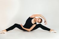 Young flexible female in black outfit and pointes stretching while practicing yoga and looking at camera in studio on white background
