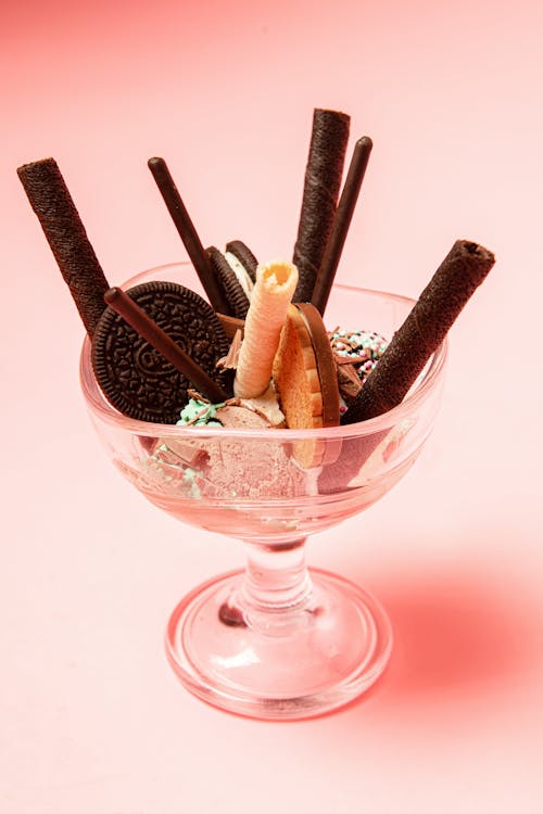 Transparent glass of tasty gelato scoops with assorted waffle rolls and crunchy biscuits on pink background