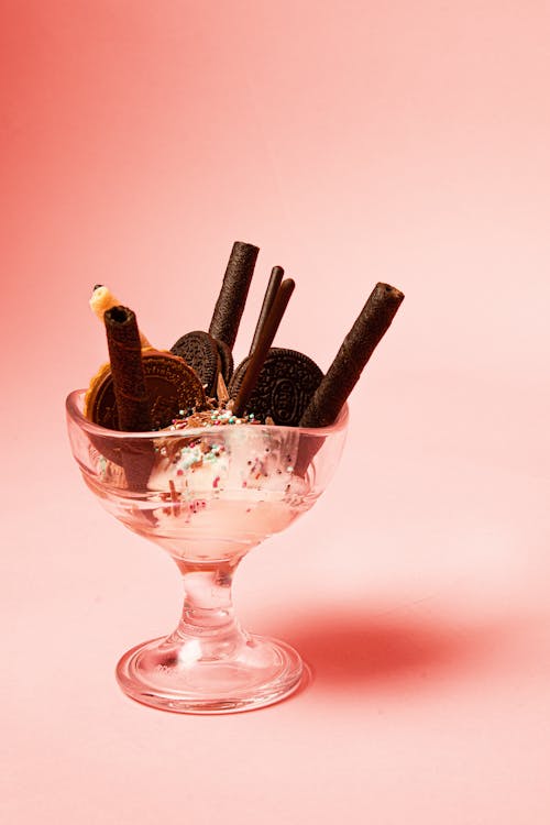 Transparent glass of yummy ice cream decorated with chocolate waffle rolls and cookies on pink background