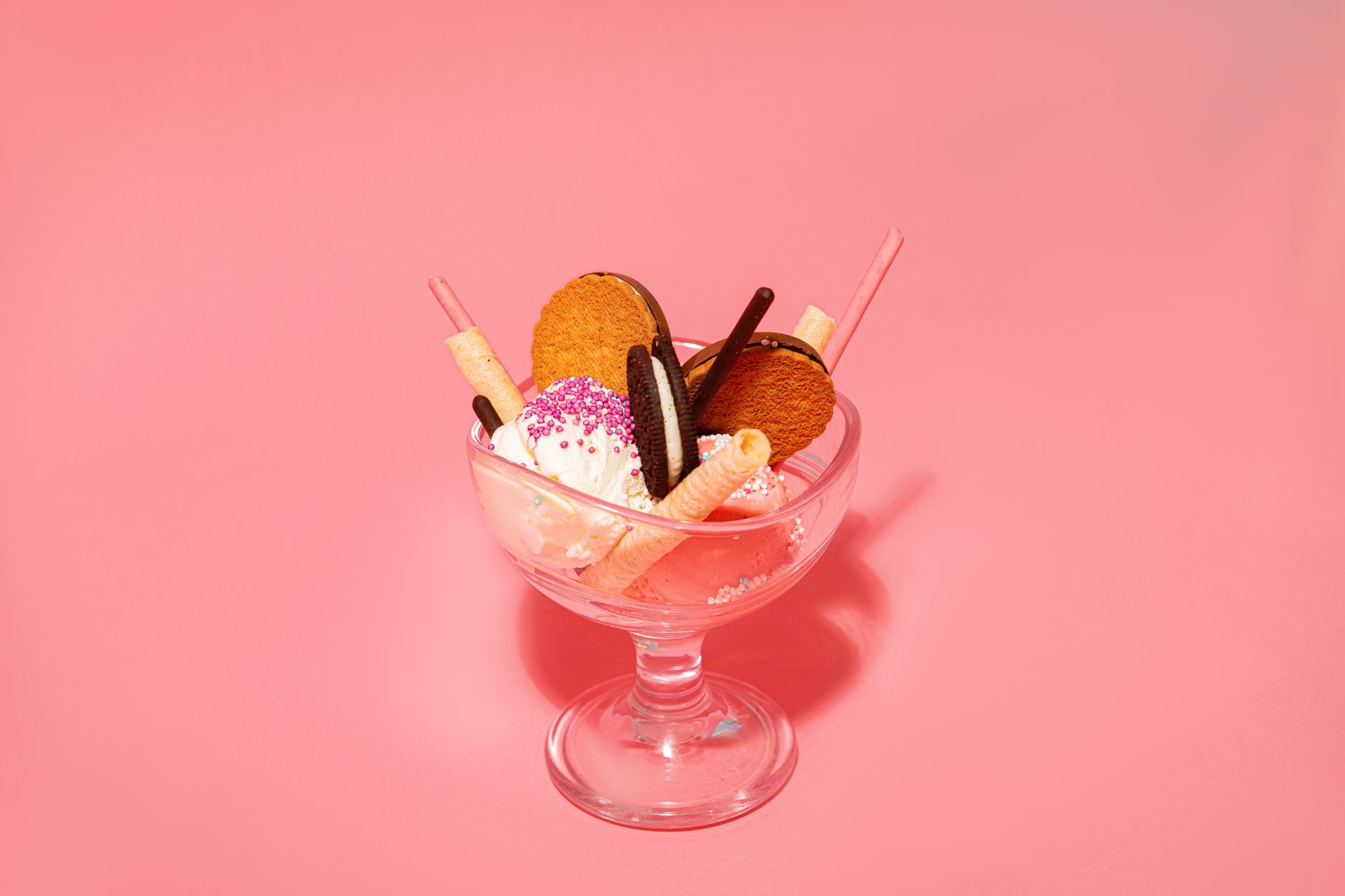 Colorful ice cream dessert with cookies and wafer sticks in a glass bowl on a pink backdrop.