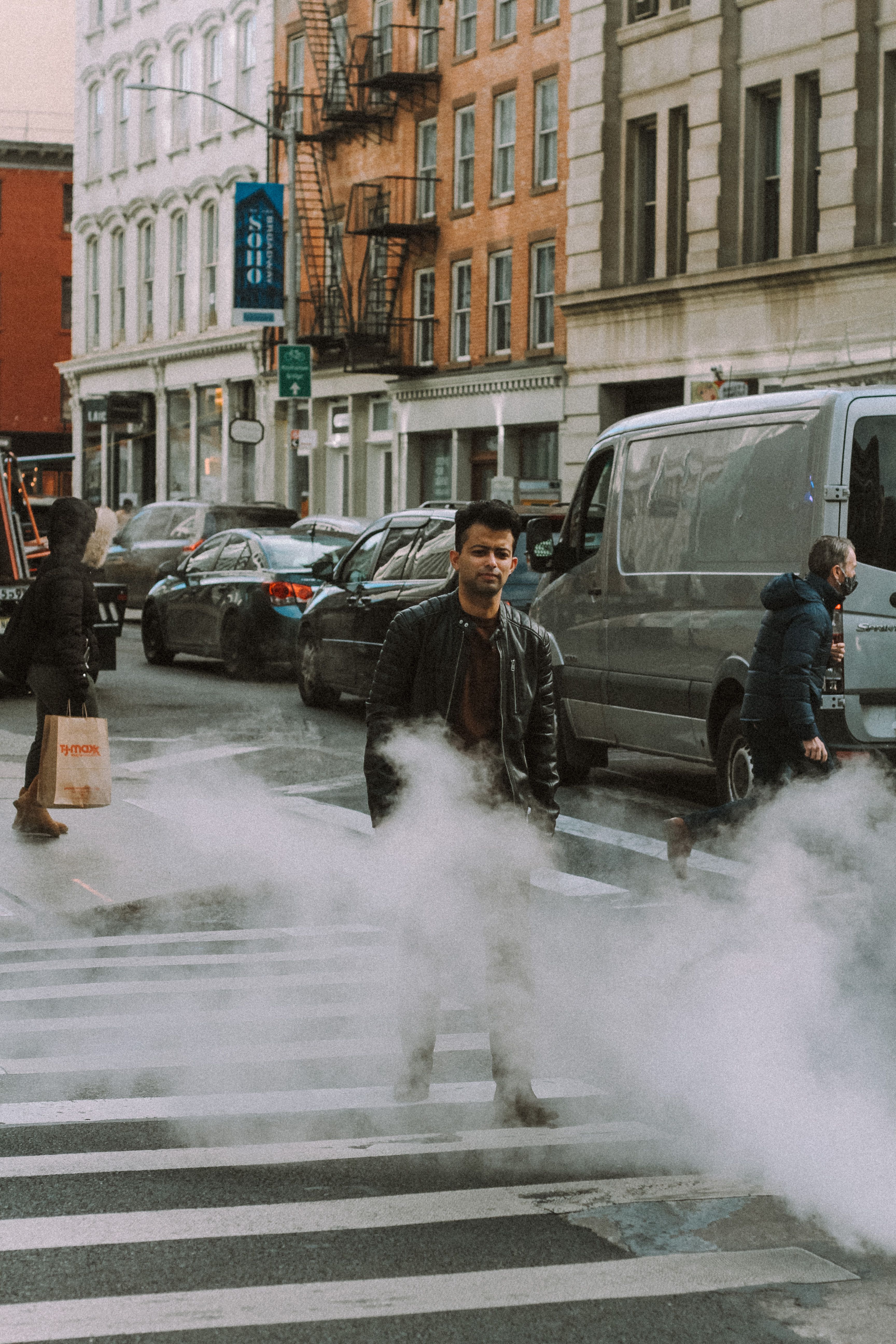 Full length of pedestrians crossing crosswalk on asphalt road with white steam near cars and buildings in town district in daytime