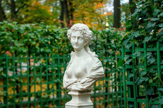 A classical white marble bust of a woman amidst a lush garden setting with a green fence.