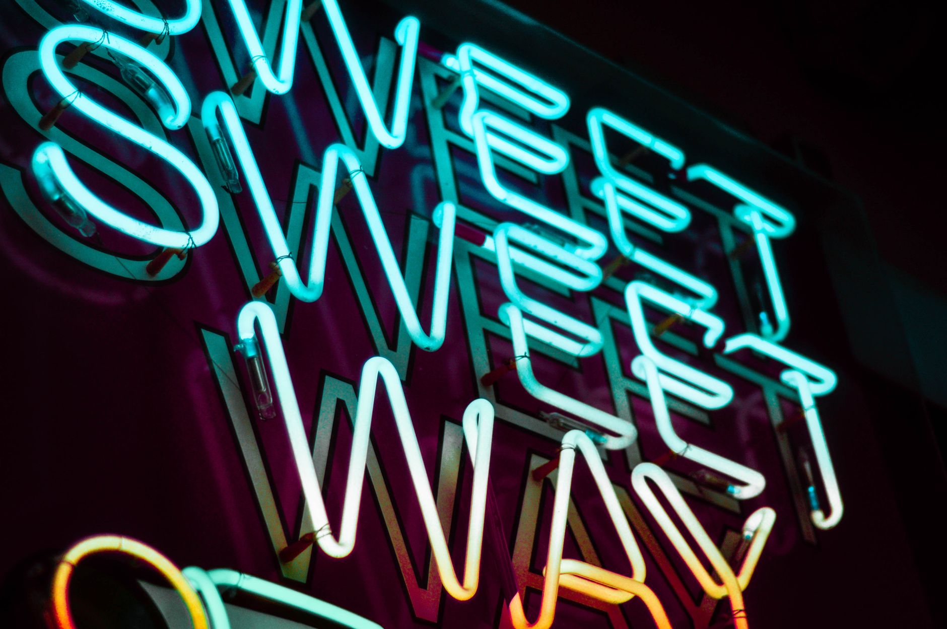 Close-up of colorful neon sign spelling 'Sweet Sweet Way' on a dark background.