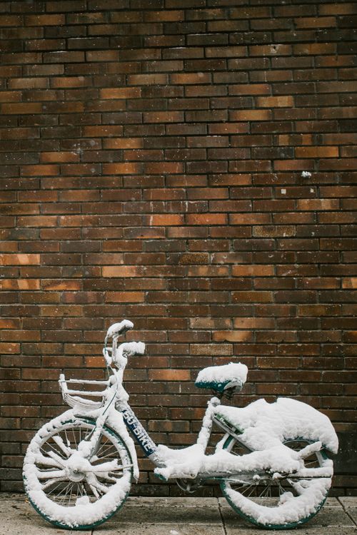 Free A solitary bicycle covered in snow leaning against a rustic brick wall, capturing winter's serene beauty. Stock Photo