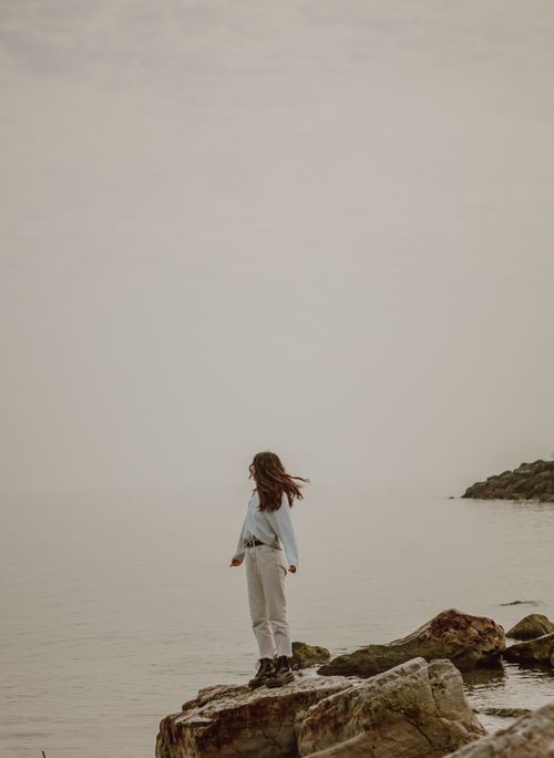 Free Faceless female traveler with flying hair in casual apparel and boots contemplating rippled ocean from rough boulder under gray sky Stock Photo
