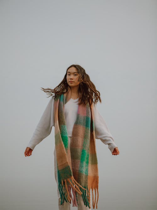Young gentle ethnic female tourist with flying hair in warm scarf looking at camera against ocean and misty sky