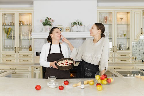Two women joyfully baking pie at home, enjoying quality family time in a cozy kitchen.