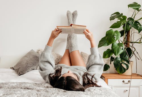 Unrecognizable female in warm clothes reading textbook while lying with crossed legs on bed in house