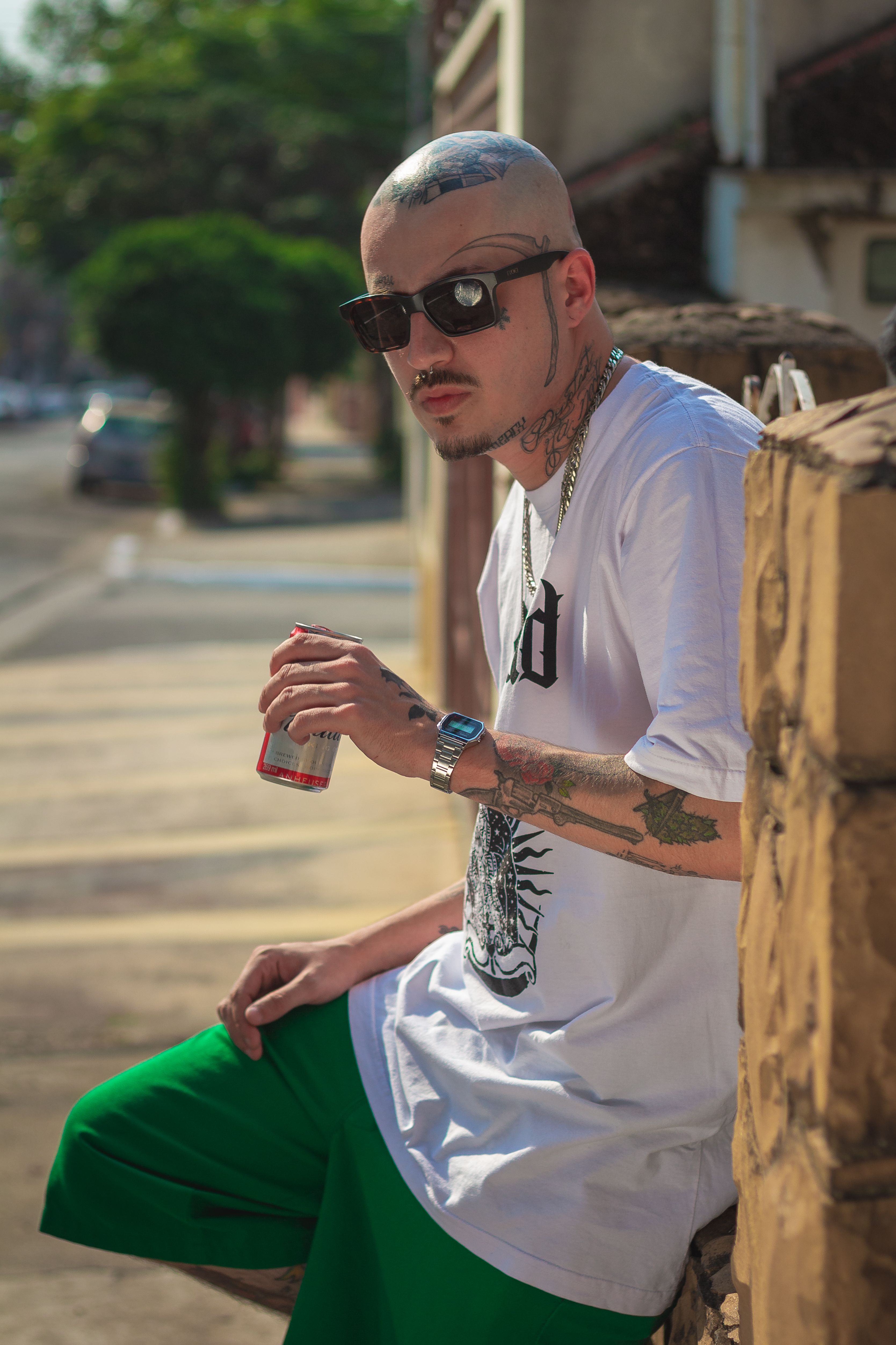 Tattooed man in sunglasses leaning on a wall holding a beverage can in a city street.