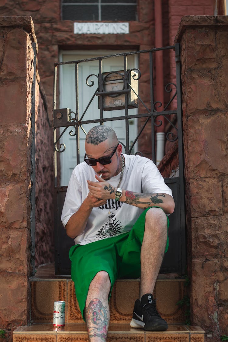 Man With Tattoos Sitting On Stairs At House Gate And Lighting Cigarette