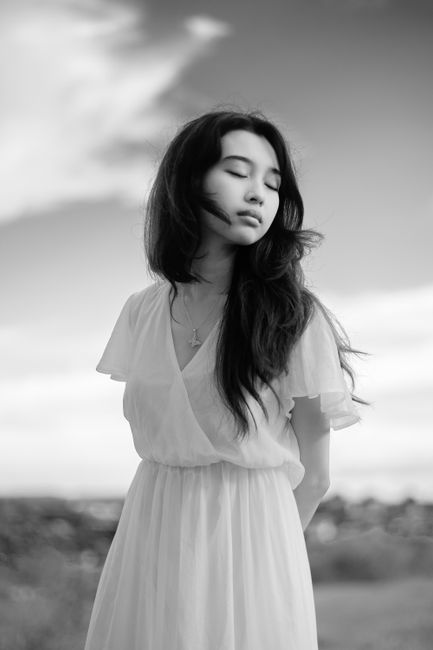 Black and white of young dreamy ethnic female with hands behind back and closed eyes under cloudy sky