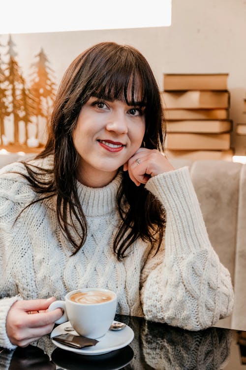 Smiling brunette in cozy white sweater sitting at table with fresh hot latte in modern cafe and looking at camera