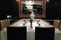 Elegant restaurant with banquet table covered with tablecloth with glasses and napkins near plates and small bottles near chairs and mirror on wall with chandelier reflection near urn for ashes