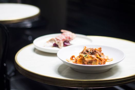 Round white table with delicious pasta with meat and herbs with sauce in bowl near plate with small roasted bird in bright restaurant