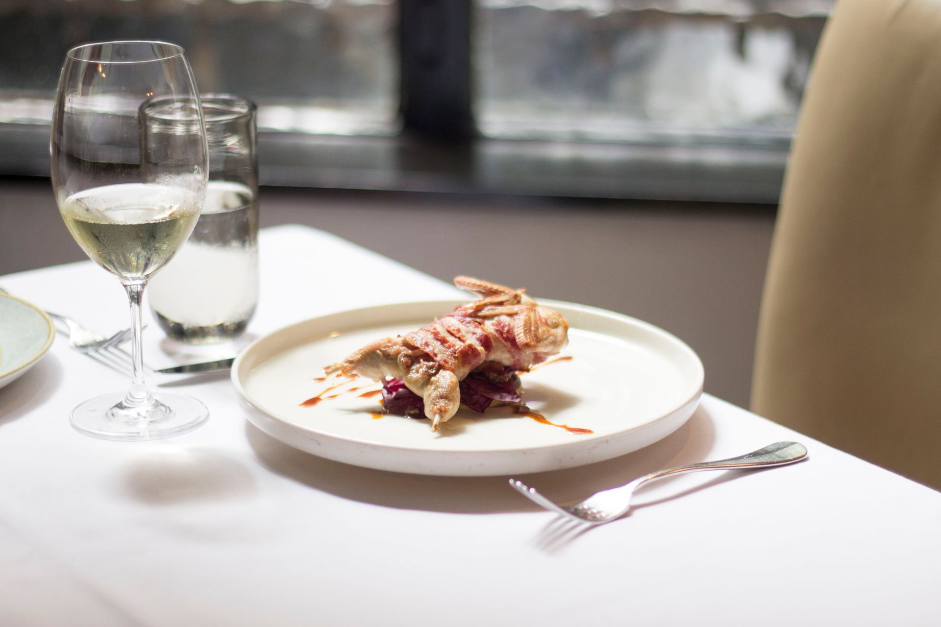 Tablecloth on table with delicious plate with small roasted bird covered in bacon with sauce near glasses with wine and water near forks and knife in light restaurant near chair