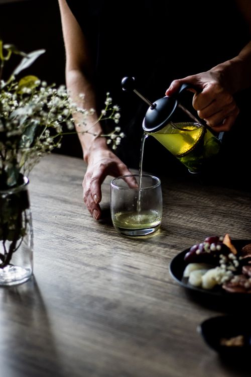 Free A person pouring green tea from a French press into a glass on a rustic wooden table. Stock Photo