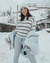 Young glad female in warm clothes looking at camera against construction while standing on snow in countryside