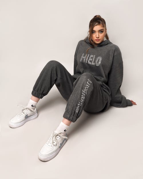 Full body of trendy female wearing trendy sportswear looking at camera while sitting on floor with bent legs on white background