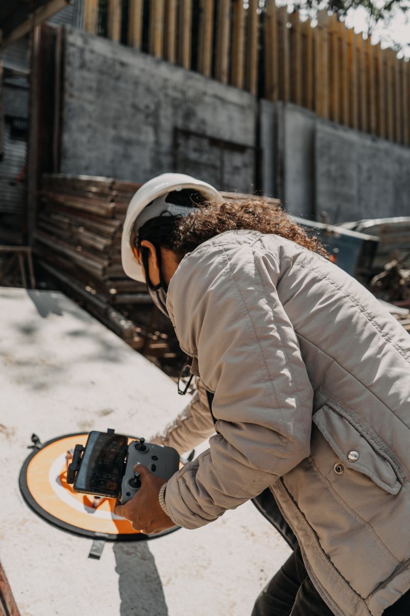 A construction worker in a hard hat uses a drone controller on a jobsite with rebar and concrete walls visible