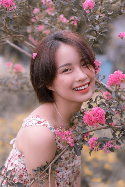 A cheerful woman with short hair surrounded by vibrant pink flowers, captured outdoors.