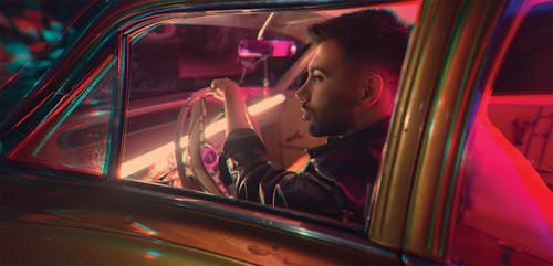 Side view of confident bearded male sitting on driver seat in vintage car with neon illumination in night time on street
