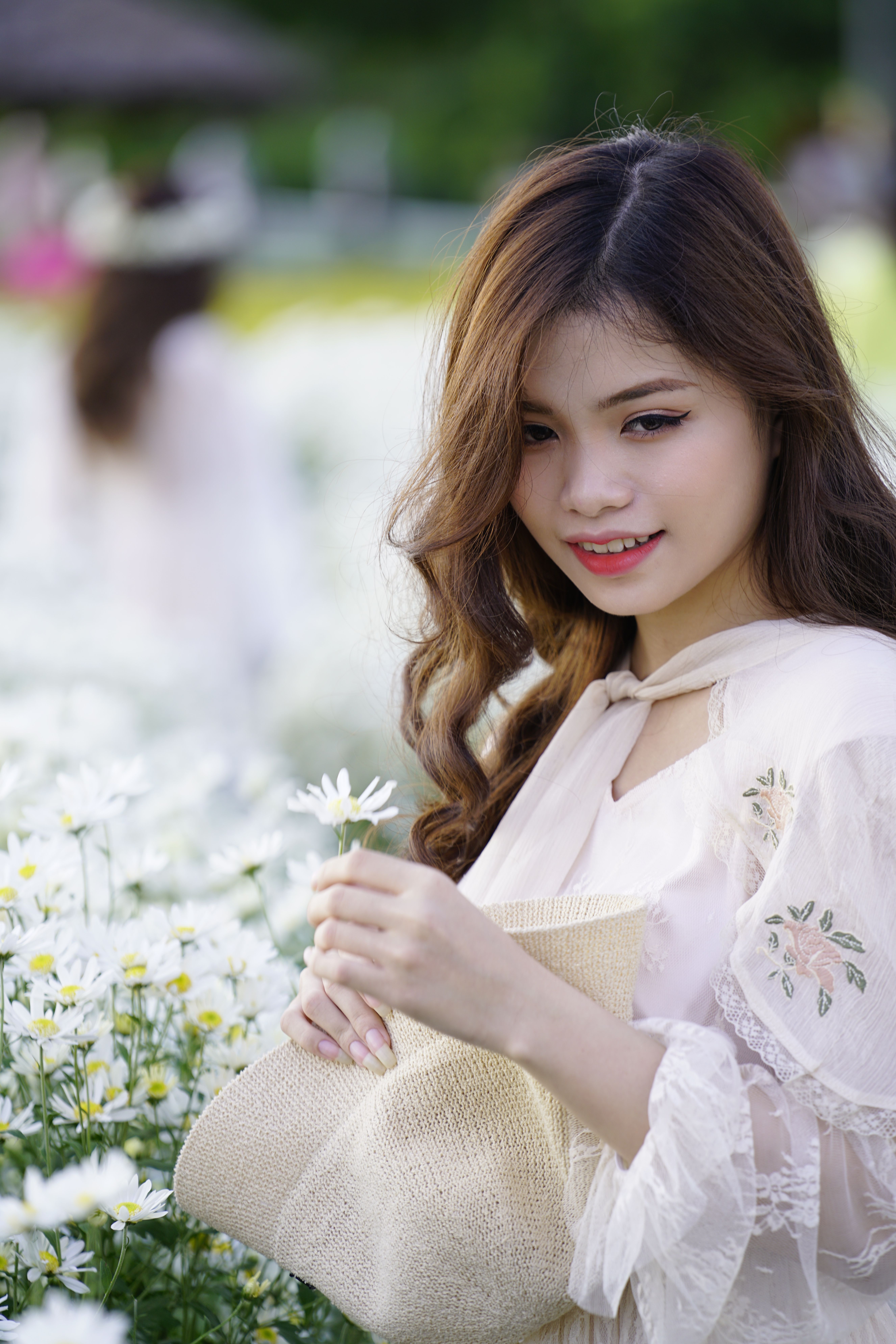 Charming Asian female adolescent with hat and blossoming flower looking down in field on blurred background