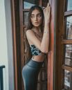 Thoughtful charming female with long hair looking over shoulder while standing on porch in doorway with raised hand on wooden brown door