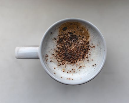 A close-up top view of a cappuccino in a white cup with coffee grinds on frothy milk, on a plain background.