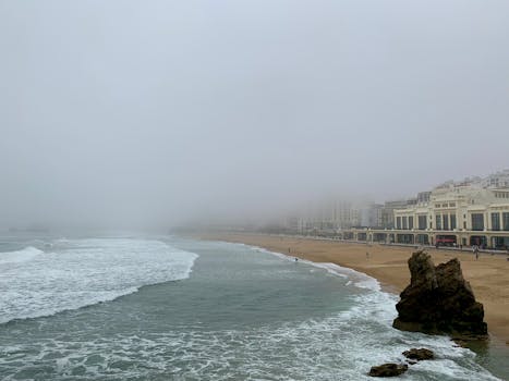 Moody seascape of Biarritz beach with fog, waves, and cityscape buildings in the distance.