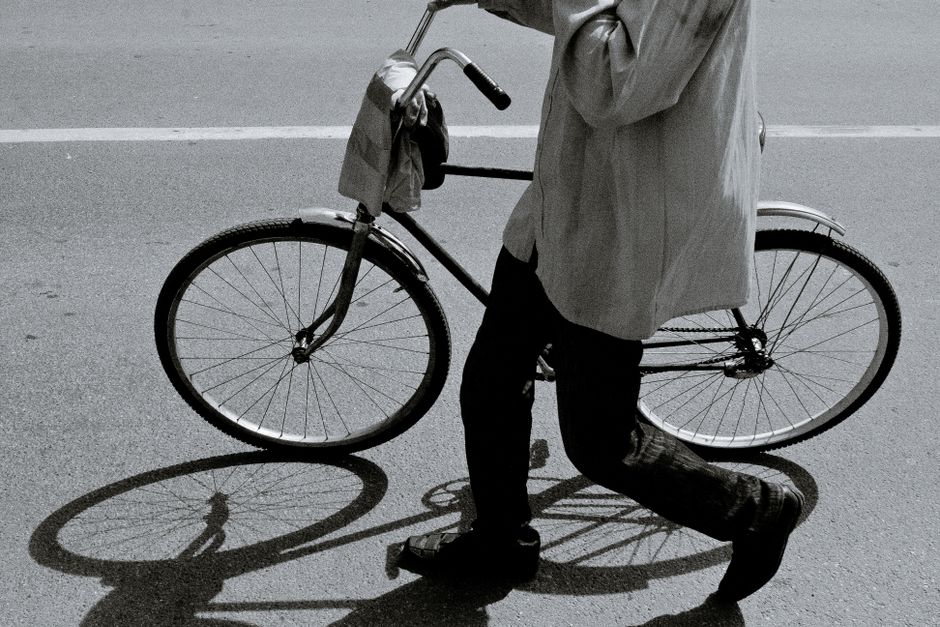 Side view of black and white crop anonymous person in stylish outfit walking on asphalt road with bicycle on sunny day
