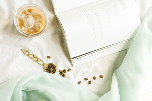 Stylish flat lay featuring iced coffee, open book, coffee beans, and a delicate cloth, perfect for lifestyle themes.