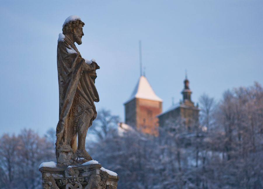 Snow-covered stone statue with Fribourg castle in the background on a serene winter day.